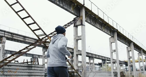 Rear of the sporty man running in a sporty clothes and a cap in the industrial facility disttrict in ruins on the cold bad weather day. Outdoor photo