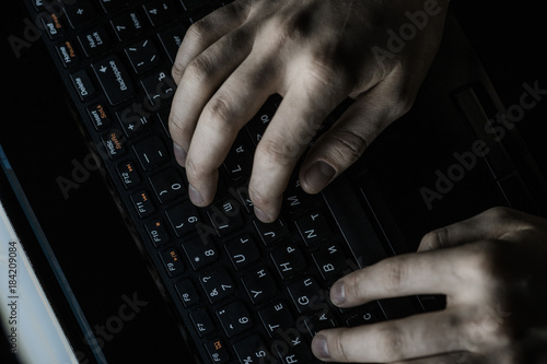 Male computer hacker wearing a hooded top leaning over a laptop in the dark. The screen light illuminates the man with a beard performing illegal activities. Black copy space on the left photo