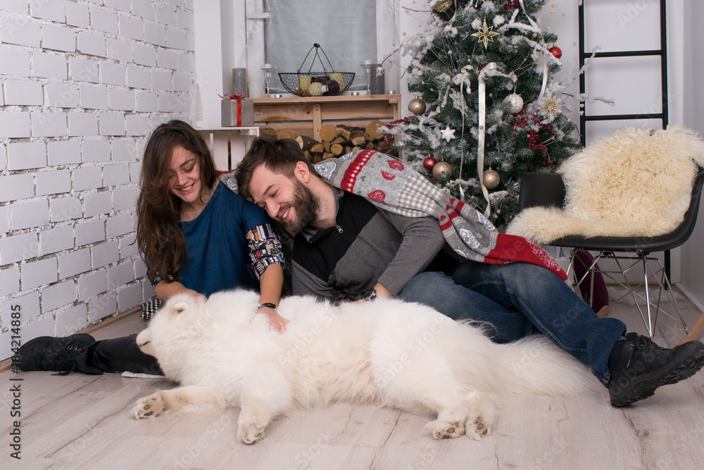 smiling girl and boy playing on the floor with his dog on the background of the New Year tree. Christmas and New Year. Samoyed and his family in a cozy room. family on Christmas smiling girl and boy playing on the floor with his dog on the background of the New Year tree. Christmas and New Year. Samoyed and his family in a cozy room. family on Christmas
