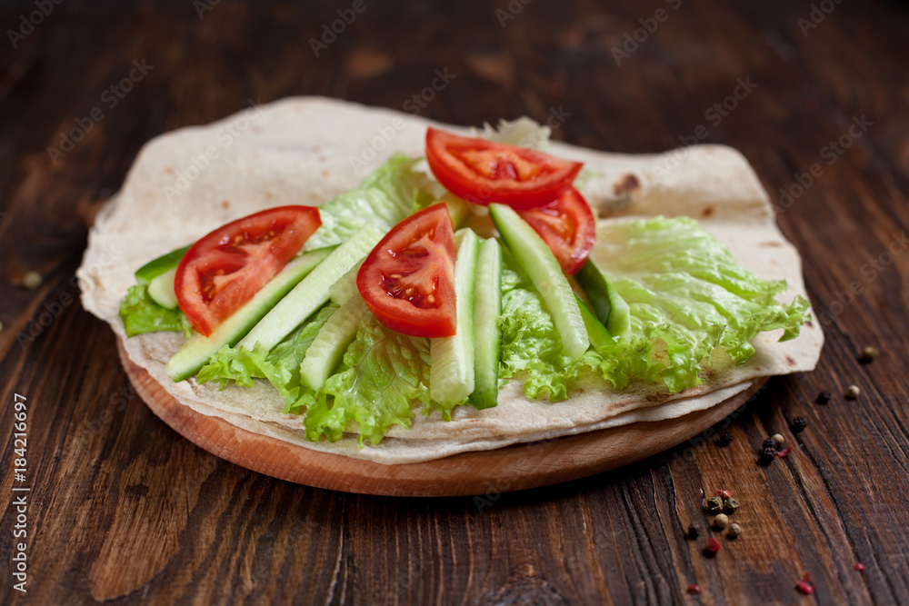 burrito with greens, cucumbers, tomatoes and sausages on a round wooden board on a wooden background burrito with greens, cucumbers, tomatoes and sausages on a round wooden board on a wooden background