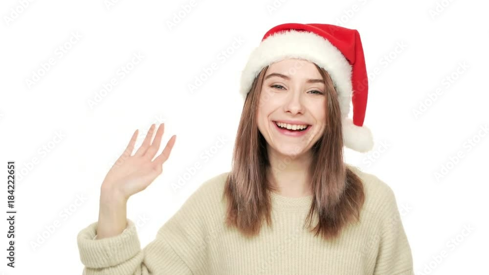 Happy portrait of attractive young woman in Santa Claus red hat greeting people waving hand enjoying favourite winter holiday over white background copy space. Concept of emotions Happy portrait of attractive young woman in Santa Claus red hat greeting people waving hand enjoying favourite winter holiday over white background copy space. Concept of emotions