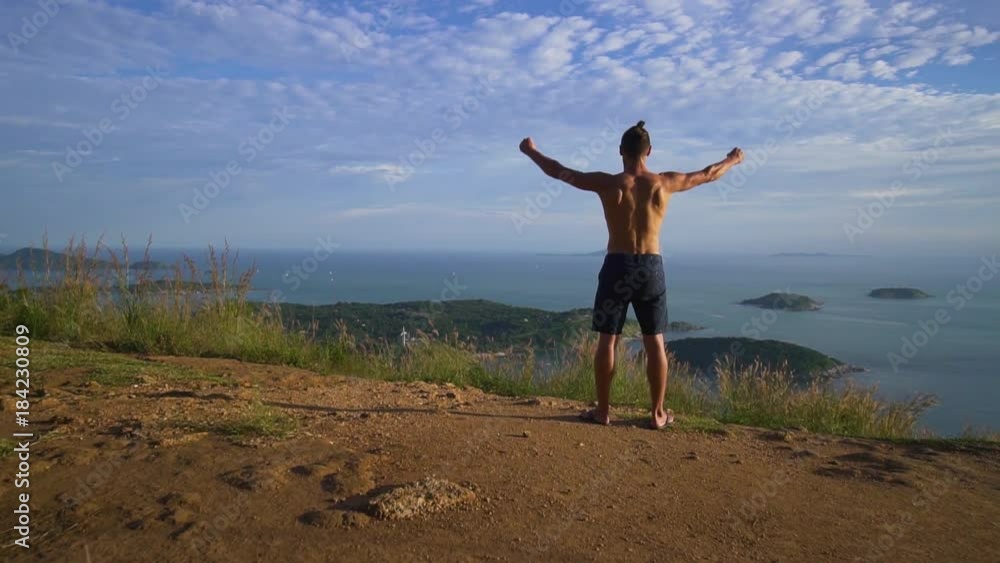 Athletic young man with bare chest raises his hands up standing on the edge of the mountain overlooking the sea and the islands. Celebrates the victory. Rear back view. Phuket. Thailand. Athletic young man with bare chest raises his hands up standing on the edge of the mountain overlooking the sea and the islands. Celebrates the victory. Rear back view. Phuket. Thailand.
