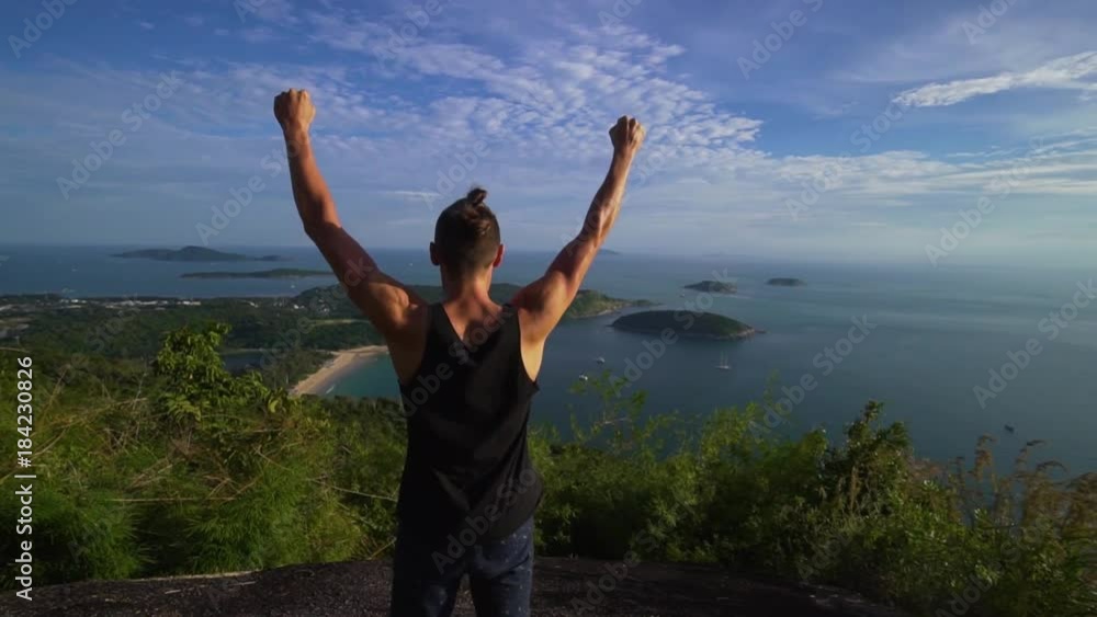 Athletic young man stands with his hands up on the edge of the mountain overlooking the sea and the islands. Celebrates the victory. Rear back view. Phuket. Thailand. Athletic young man stands with his hands up on the edge of the mountain overlooking the sea and the islands. Celebrates the victory. Rear back view. Phuket. Thailand.