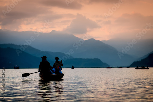 Silhouettes of people on boats at sunset on Phewa lake. Vanilla sky and mountain background.