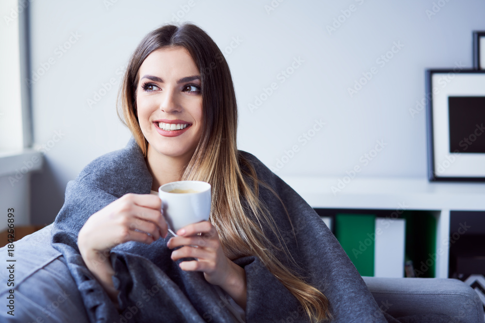 Cheerful young woman drinking coffee sitting under blanket on sofa at home - obrazy, fototapety, plakaty 