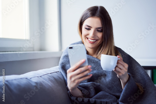 Smiling young woman sitting under blanket using phone and having tea at home