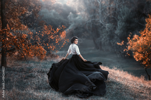 girl in black and white vintage dress is running looking around. Silk fabric skirt train waving fly in wind motion. Artistic Photography. Medieval fantasy woman walk in autumn nature, tree orange leaf photo
