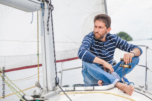 man sits on bow of sailing yacht and holds binoculars