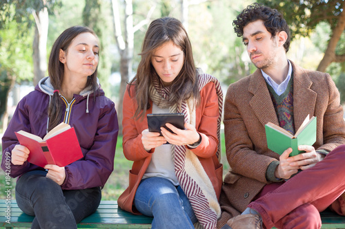 One Woman Sitting on a Bench in a Park Reading an E-Book with Two People with Paper Books Peeping