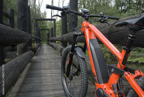 Panorama: Ebike E-bike electric bicycle orange, detail from back, leaning on an old dark wooden bridge, in the woods, beneath which flows a small light green mountain river, Ossola, Alps, Italy