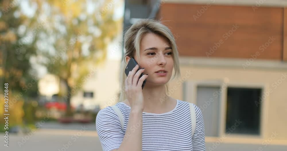 Beautiful young female having conversation on mobile phone during walk on street, modern architecture building background Beautiful young female having conversation on mobile phone during walk on street, modern architecture building background