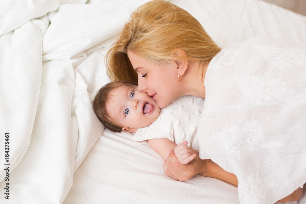Portrait of young mother with blond hair with her sweet 3 month old baby girl in white wear having fun in the bedroom at morning, loving happy family concept, top view Portrait of young mother with blond hair with her sweet 3 month old baby girl in white wear having fun in the bedroom at morning, loving happy family concept, top view