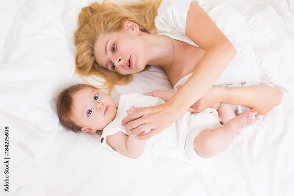 Portrait of young mother with blond hair with her sweet 3 month old baby girl in white wear having fun in the bedroom at morning, loving happy family concept, top view Portrait of young mother with blond hair with her sweet 3 month old baby girl in white wear having fun in the bedroom at morning, loving happy family concept, top view