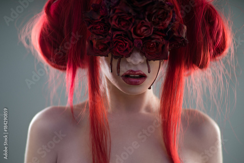 Portrait of red haired woman with flower mask and blood on face posing at camera on grey background