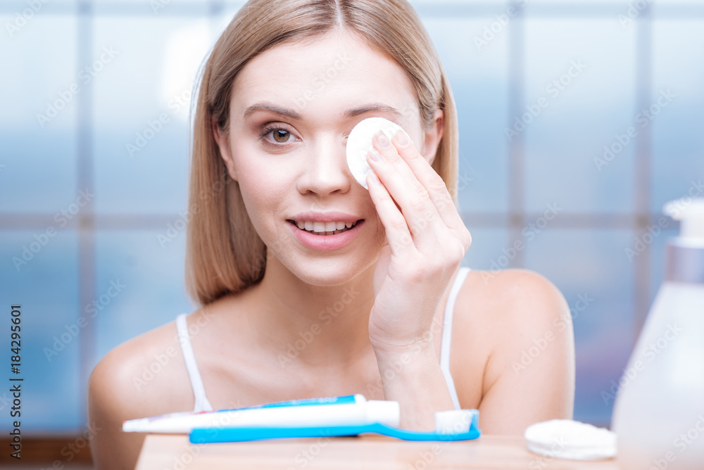 After long day. Pleasant young woman in a white tank top standing in the bathroom and removing the make up from her eyes using a cotton pad After long day. Pleasant young woman in a white tank top standing in the bathroom and removing the make up from her eyes using a cotton pad