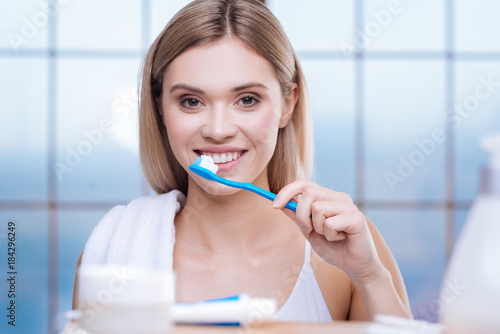 Clean teeth. Pretty fair-haired young woman standing in the bathroom and cleaning her teeth while smiling brightly at her reflection