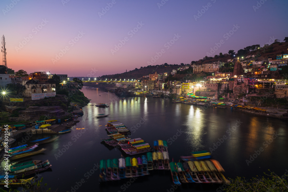 Omkareshwar cityscape at dusk, India, sacred hindu temple. Holy Narmada River, boats floating. Travel destination for tourists and pilgrims.