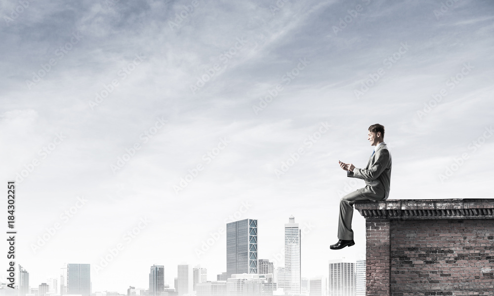 Man on roof edge send message with smartphone and cityscape at b Man on roof edge send message with smartphone and cityscape at b