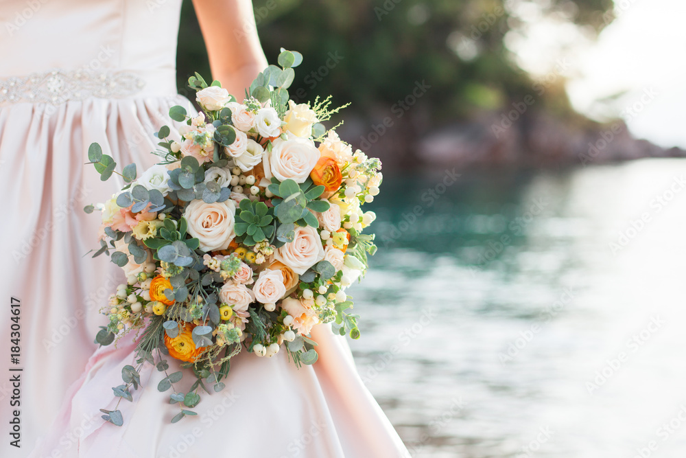 Bride holds wedding bouquet from roses and stands on background of sea and beach after wedding ceremony. Bride is dressed in cream wedding dress. Bride holds wedding bouquet from roses and stands on background of sea and beach after wedding ceremony. Bride is dressed in cream wedding dress.