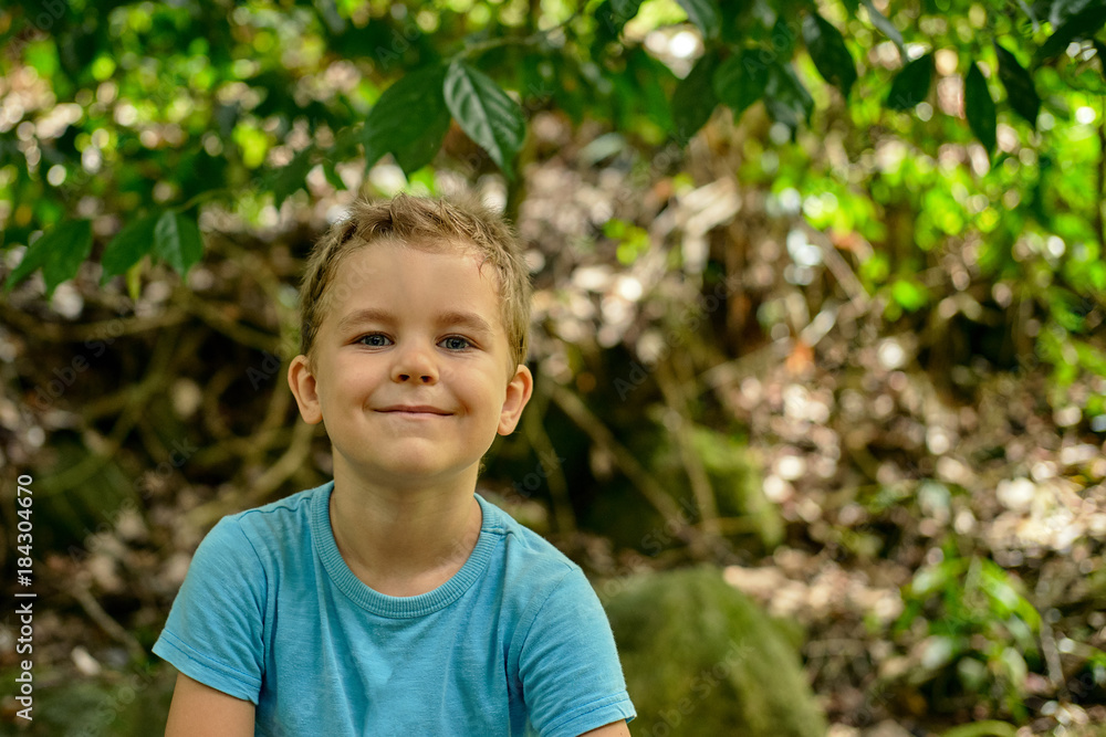 Portrait of a boy 6 years old cheekily smiles against a background of green foliage outdoor, blue shirt, blonde hair, Portrait of a boy 6 years old cheekily smiles against a background of green foliage outdoor, blue shirt, blonde hair,