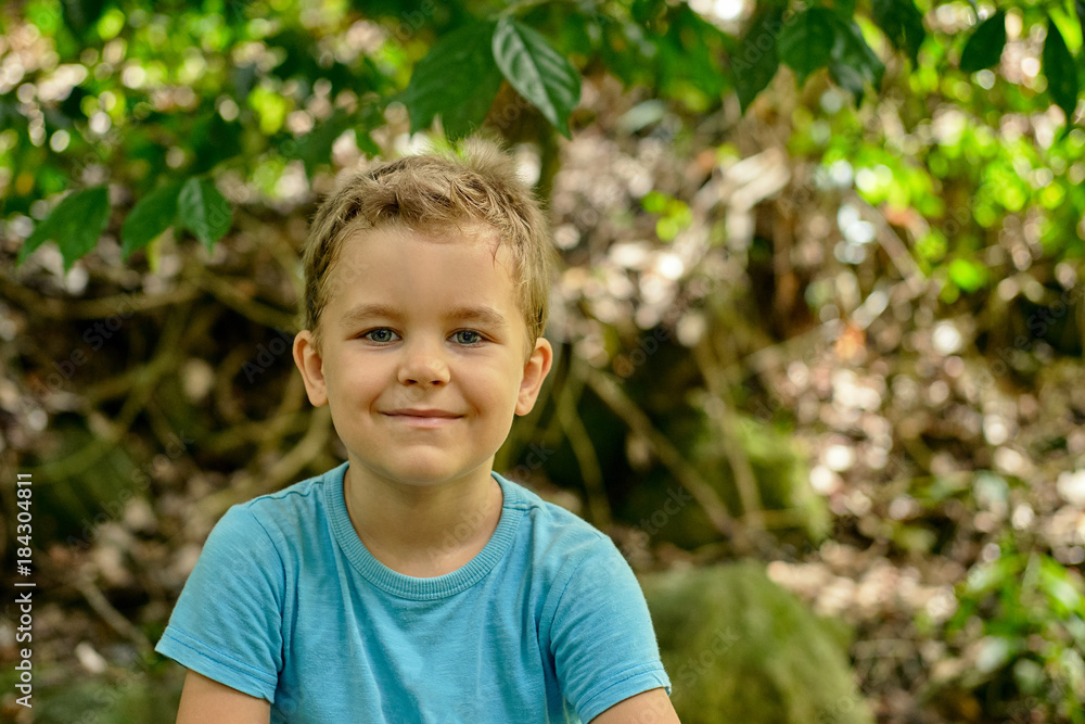 Portrait of a boy 6 years old cheekily smiles against a background of green foliage outdoor, blue shirt, blonde hair, Portrait of a boy 6 years old cheekily smiles against a background of green foliage outdoor, blue shirt, blonde hair,