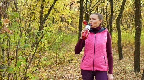 Slow motion. Beautiful fitness sport girl in sportswear drinks water or isotonic drink from a sports water bottle in the autumn park. photo