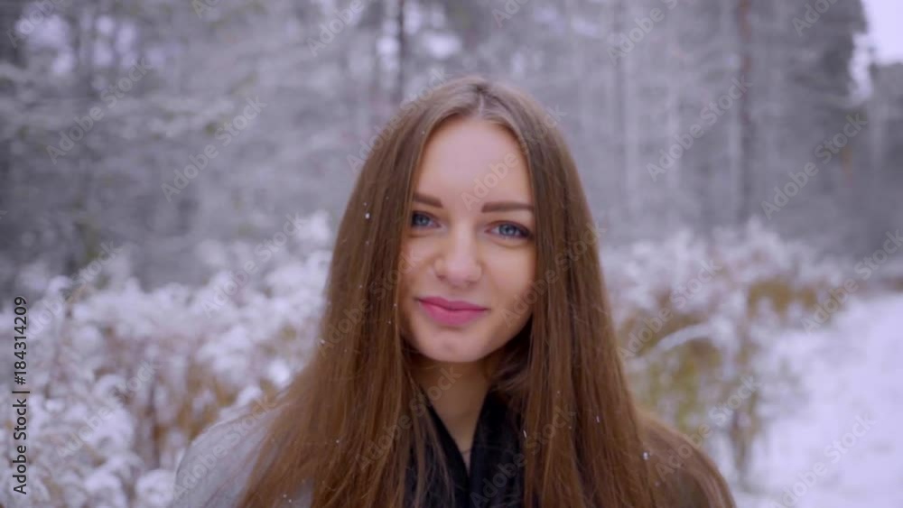 Close up Portrait of stylish happy young beautiful woman outdoors in a winter park over snowy background. looking to the camera. It is snowing. beautiful young woman is walking in a winter park. Slow Close up Portrait of stylish happy young beautiful woman outdoors in a winter park over snowy background. looking to the camera. It is snowing. beautiful young woman is walking in a winter park. Slow