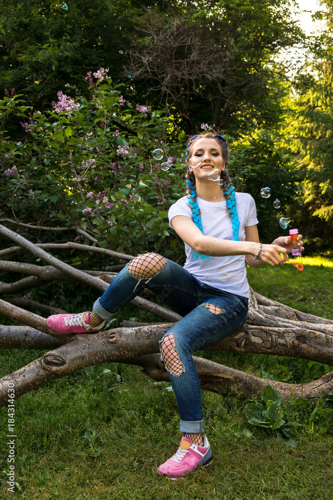 Young girl with two blue braids sits in a park on a tree and blows up soap bubbles, has fun