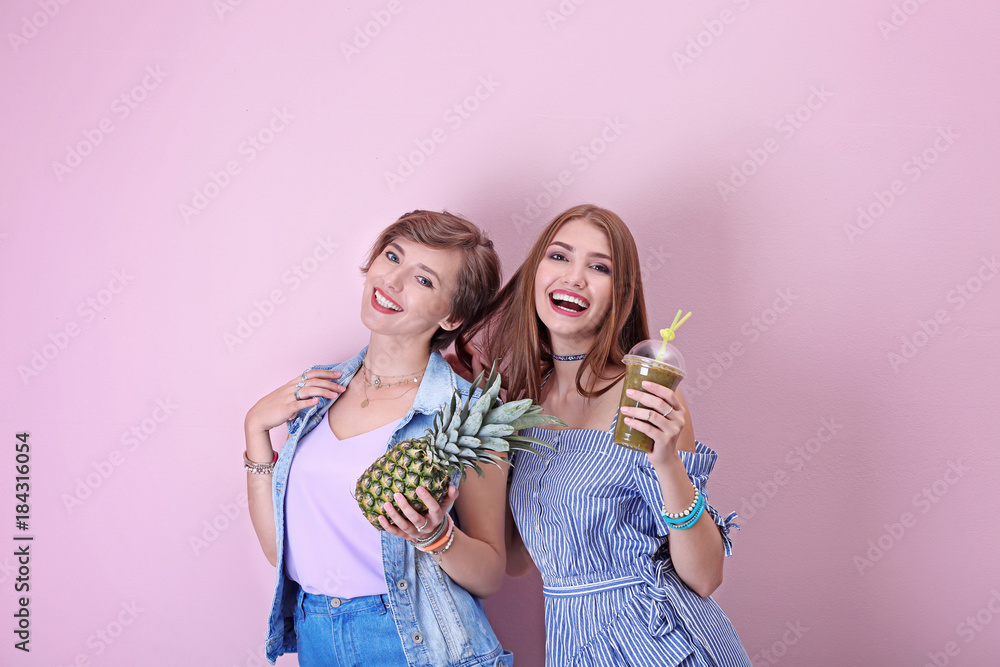Attractive hipster girls posing with pineapple and smoothie on color background Attractive hipster girls posing with pineapple and smoothie on color background