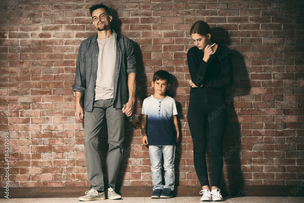Man and woman standing near their little son with bottle of alcohol and cigarette against brick wall background Man and woman standing near their little son with bottle of alcohol and cigarette against brick wall background