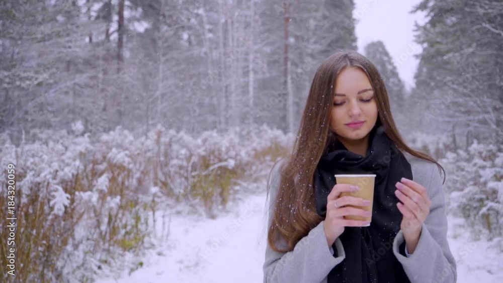 Beautiful young woman with disposable coffee cup is drinking tea or coffee outdoors in a winter park. Slow motion. Portrait of stylish young beautiful woman walking in a winter park over snowy Beautiful young woman with disposable coffee cup is drinking tea or coffee outdoors in a winter park. Slow motion. Portrait of stylish young beautiful woman walking in a winter park over snowy