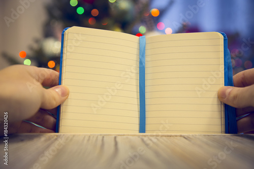 Hands holding an opened notebook on a wooden table against decorated Christmas tree with colorful fairy lights. Blurred bokeh background. Close up.Mock up for display of product © 976photo