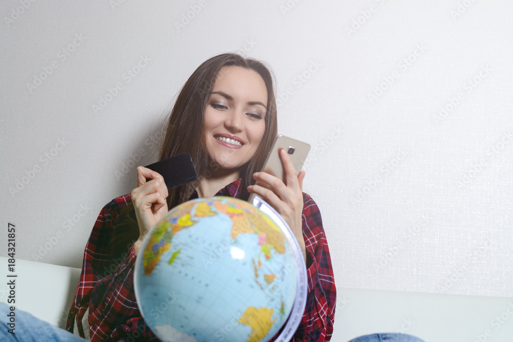 Happy woman with credit card and phone in hand, smiling sitting on sofa with globe. The joy of buying tickets for travel Happy woman with credit card and phone in hand, smiling sitting on sofa with globe. The joy of buying tickets for travel