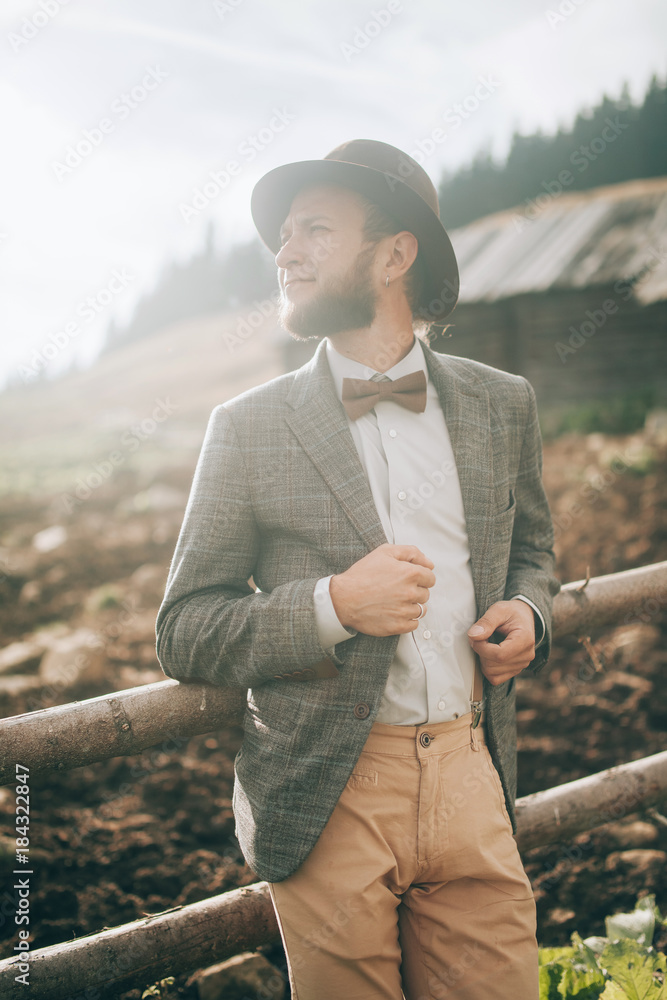 Young man in stylish clothes and hat stands on background of old wooden hut and fence. Closeup. Young man in stylish clothes and hat stands on background of old wooden hut and fence. Closeup.