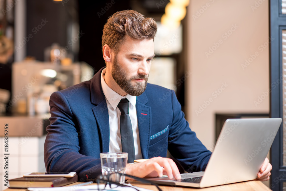 Portrait of a handsome businessman stricly dressed in the suit working with laptop at the modern cafe interior