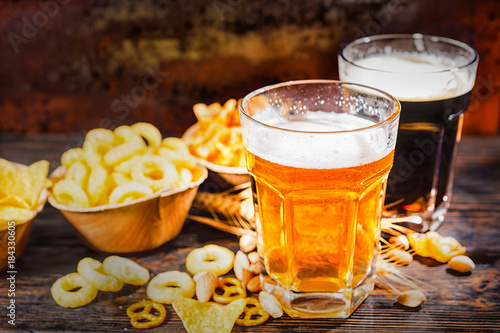 Two glasses with light and dark beer near plates with chips and scattered snacks on dark wooden desk