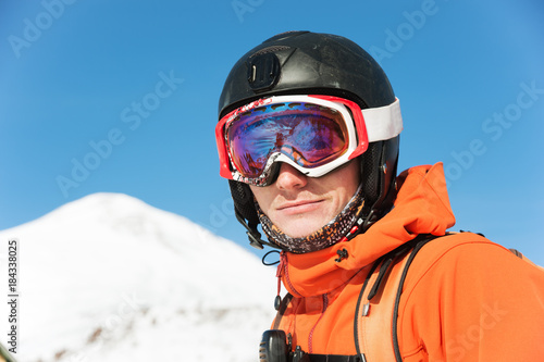 Portrait of a skier in an orange overall with a backpack on his back in a helmet stands against the background of a beautiful Caucasian mountain landscape
