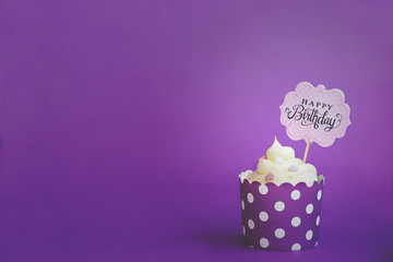 Vanilla cupcake with small decorative hearts and happy birthday sign, in violet baking paper cup, against violet background
