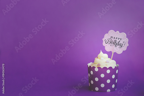 Vanilla cupcake with small decorative hearts and happy birthday sign, in violet baking paper cup, against violet background