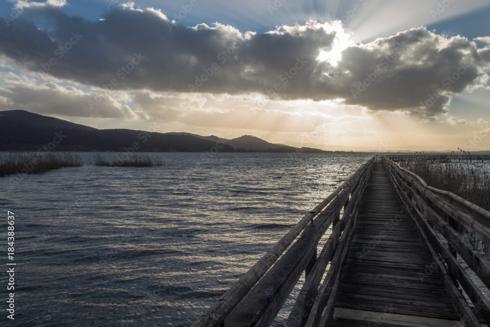 First person view of a long pier on Trasimeno lake, (Umbria), with beautiful sun rays coming out through some clouds