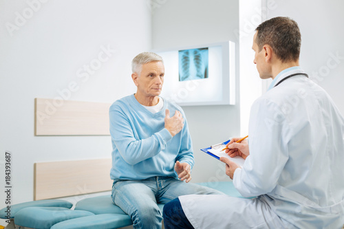 Heart problems. Nice pleasant serious man looking at his doctor and pointing at the heart while telling about his health problem