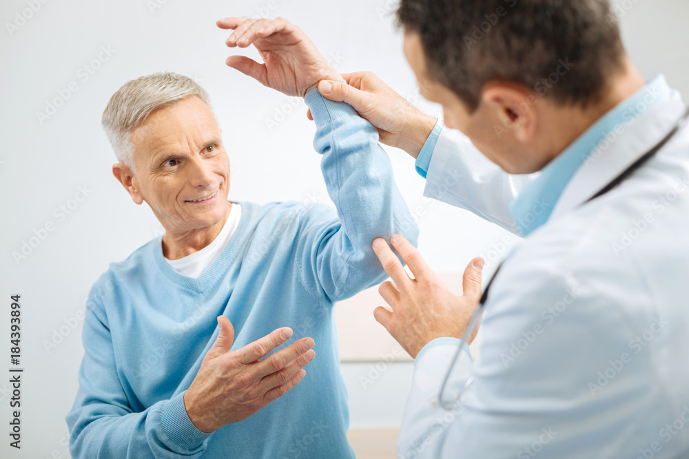 Fototapeta premium Caring about health. Nice cheerful senior man looking at his therapist and raising his hand up while smiling