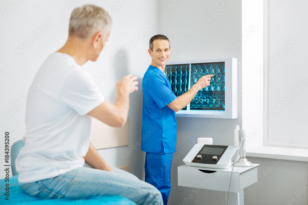 Diagnosis explanation. Cheerful delighted nice doctor standing near the X ray images and pointing at them while talking to his patient Diagnosis explanation. Cheerful delighted nice doctor standing near the X ray images and pointing at them while talking to his patient