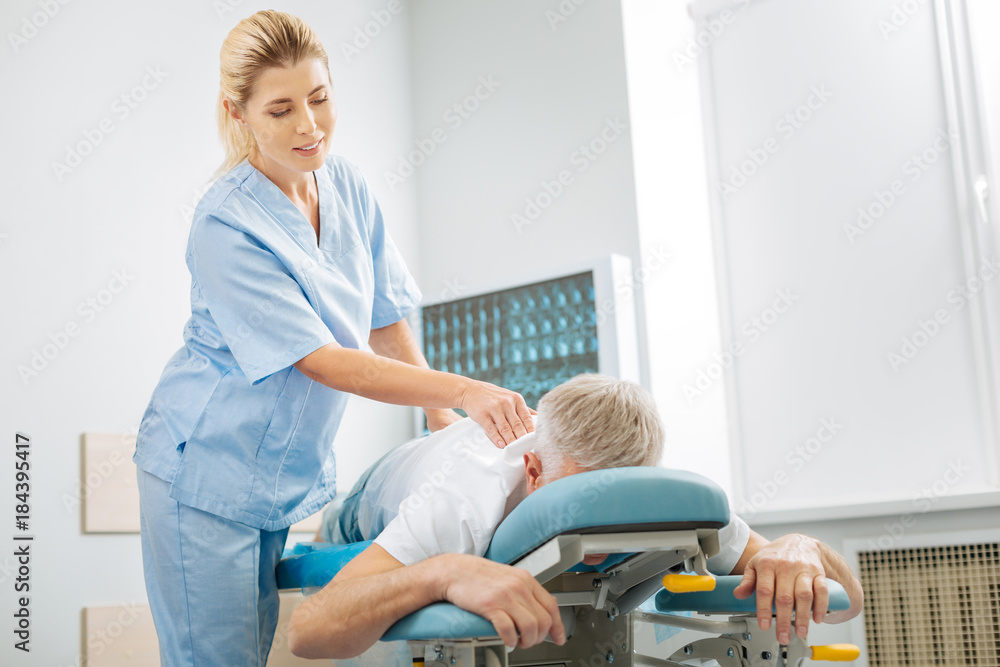 Professional medical checkup. Positive skillful female doctor standing near her patient and looking at him while doing a medical checkup Professional medical checkup. Positive skillful female doctor standing near her patient and looking at him while doing a medical checkup