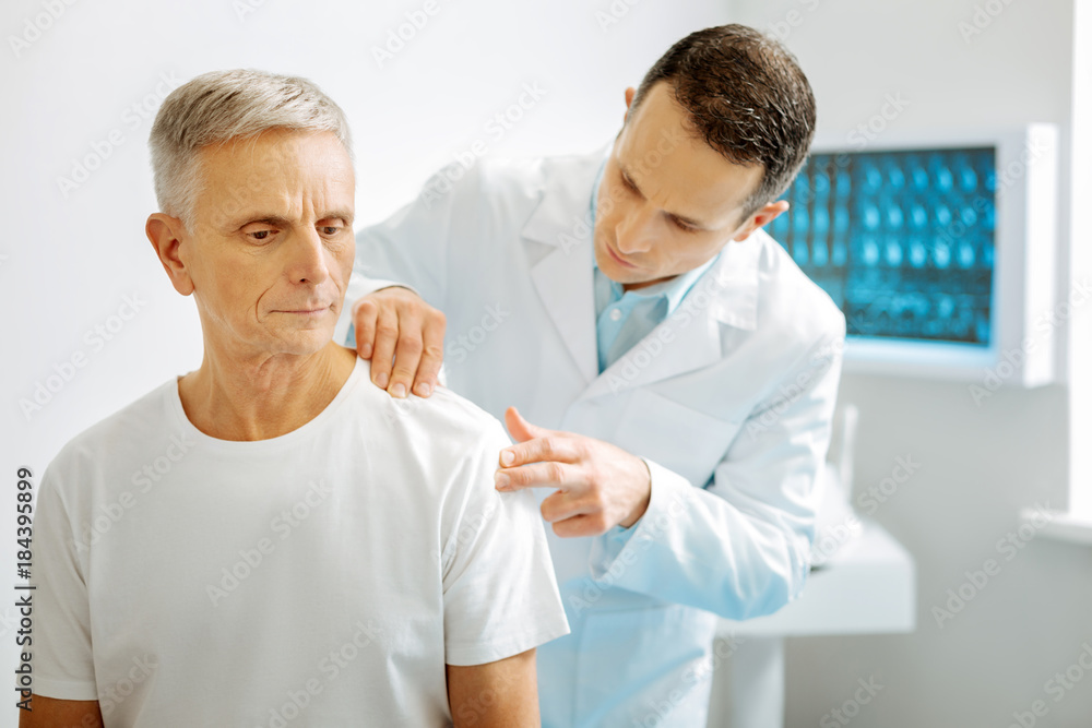 Joint ache. Serious nice smart doctor standing behind his patient and looking at his patients shoulder while checking joints Joint ache. Serious nice smart doctor standing behind his patient and looking at his patients shoulder while checking joints