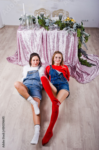 Two fun beautiful girls friends wear in overalls jeans shorts and gaiters against table with new year decoration. © AS Photo Family