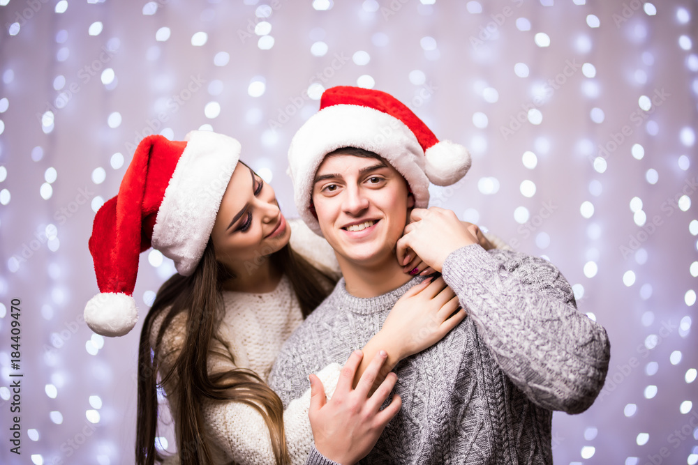 Cheerful attractive couple in winter clothes and santa hats hugging and kissing on bokeh light background Cheerful attractive couple in winter clothes and santa hats hugging and kissing on bokeh light background