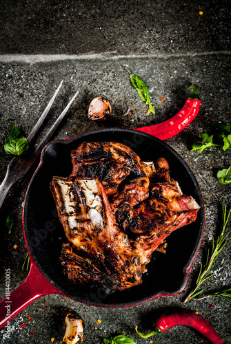 Raw fresh meat, roasted or grilled lamb or beef ribs with red tomato sauce, hot pepper, garlic and spices in skillet on dark stone background, copy space top view