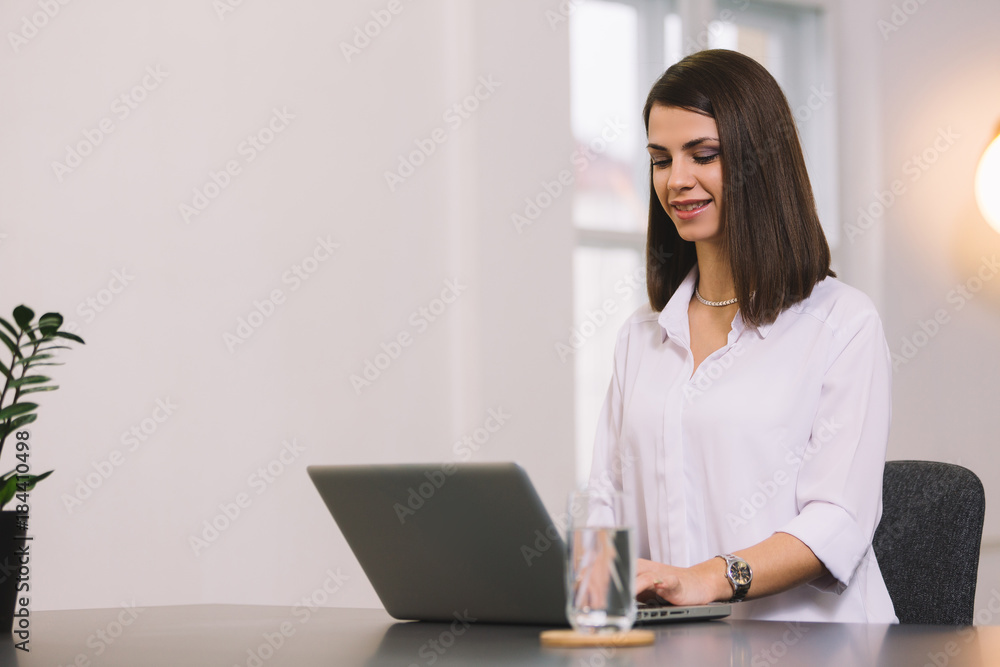 Attractive cheerful young business woman working on laptop and smiling while sitting at her desk in bright office Attractive cheerful young business woman working on laptop and smiling while sitting at her desk in bright office