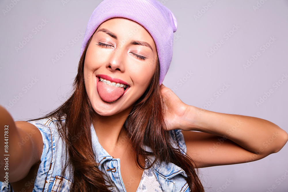 Portrait of beautiful cute brunette woman model in casual summer jeans clothes with no makeup in purple beanie making selfie photo on smartphone isolated on gray. Showing her tongue Portrait of beautiful cute brunette woman model in casual summer jeans clothes with no makeup in purple beanie making selfie photo on smartphone isolated on gray. Showing her tongue